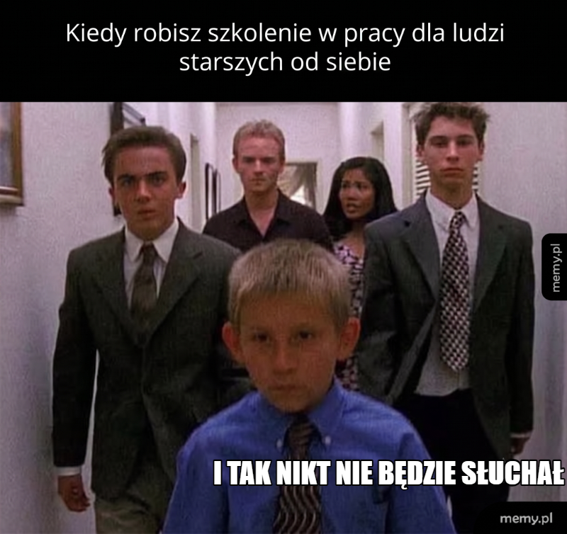 Grupa ludzi w biurze, młody chłopiec na pierwszym planie z poważnym wyrazem twarzy.